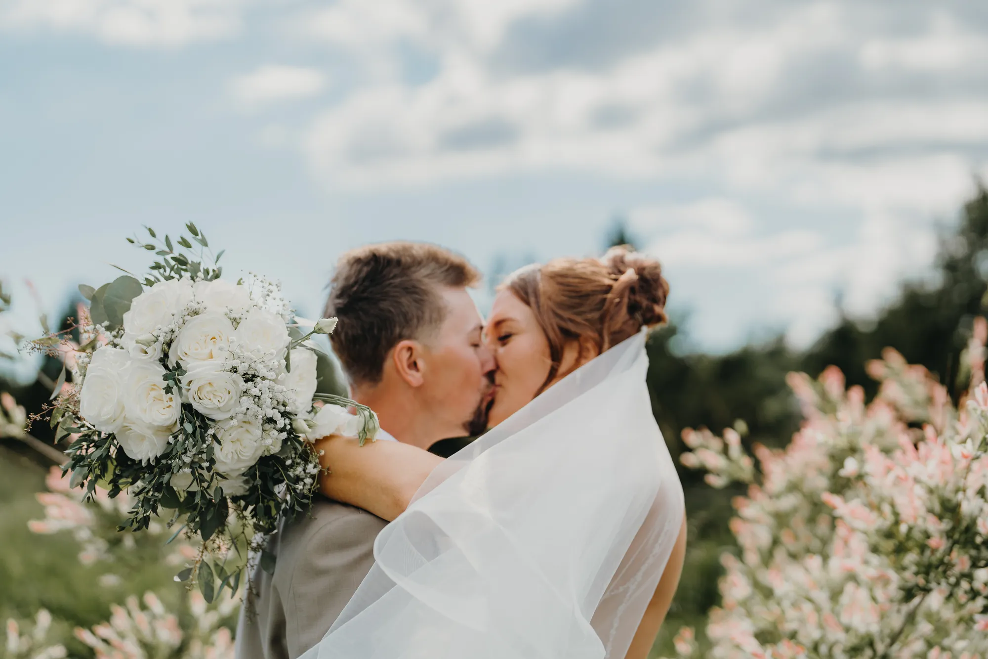 Bride and groom kissing among flowering bushes with bouquet in foreground